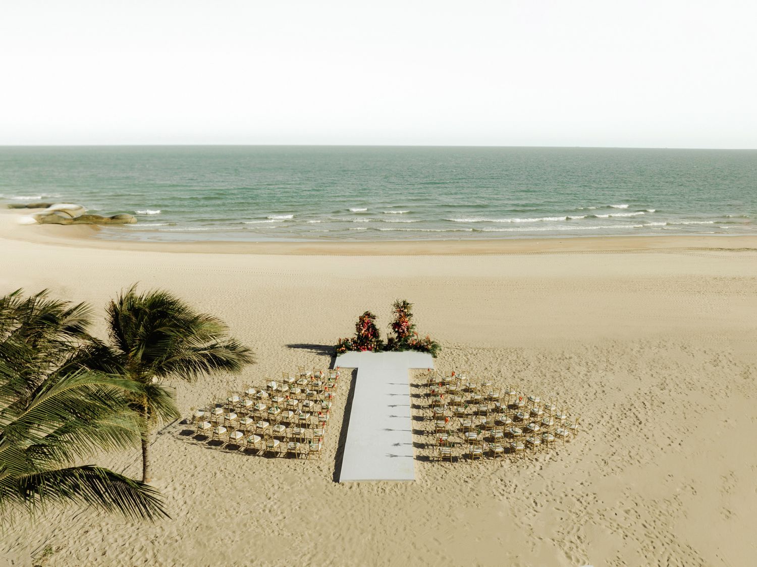 Aerial view of an elegant beach wedding setup with white chairs on sandy shore beside turquoise ocean waters.