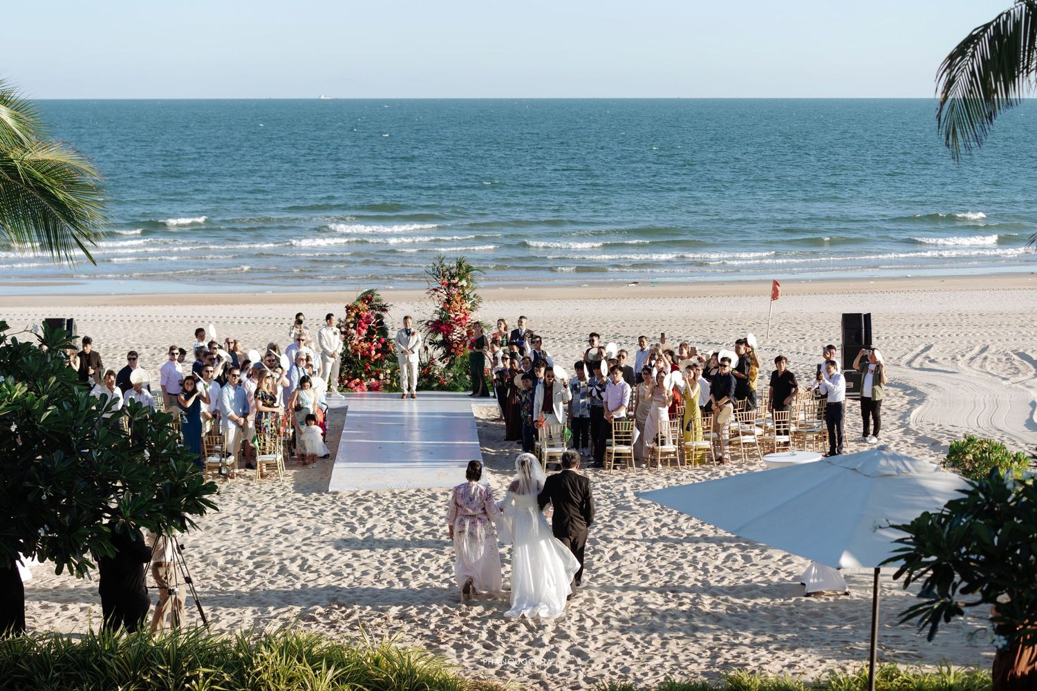 Aerial view of a romantic beach wedding ceremony with guests gathered around a floral altar by the ocean.