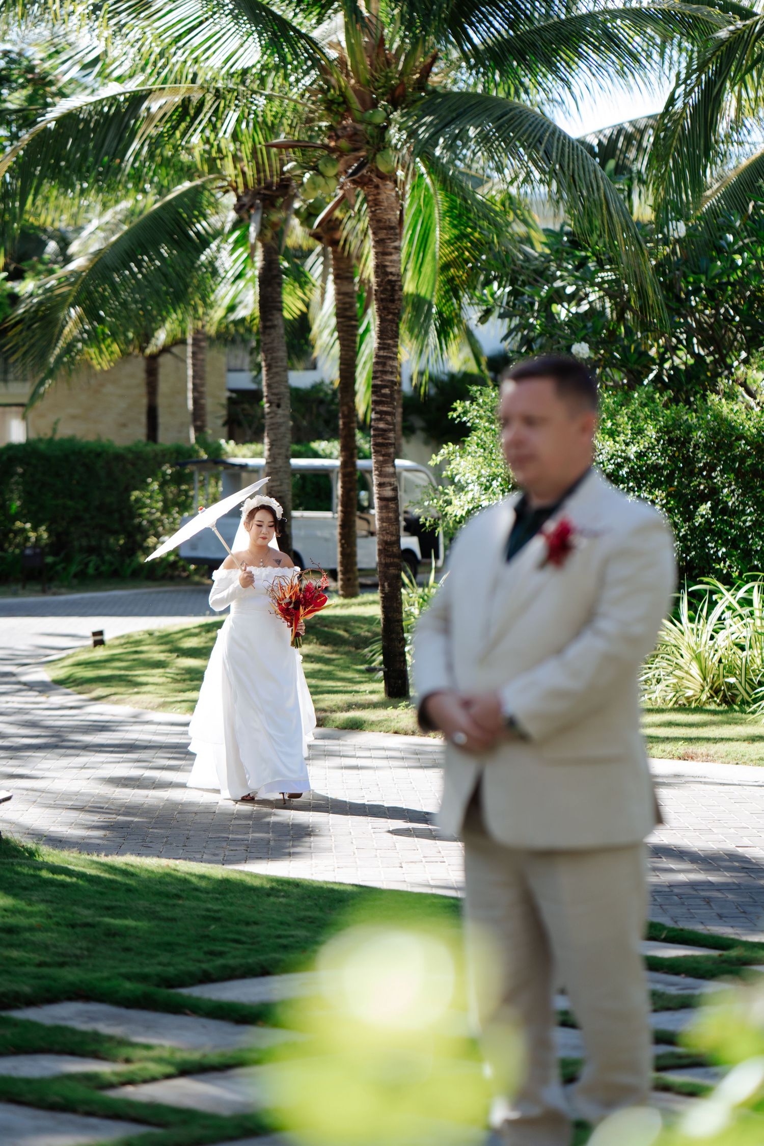 Bride in white gown walks toward groom in beige suit amid tropical palm trees during outdoor wedding ceremony.