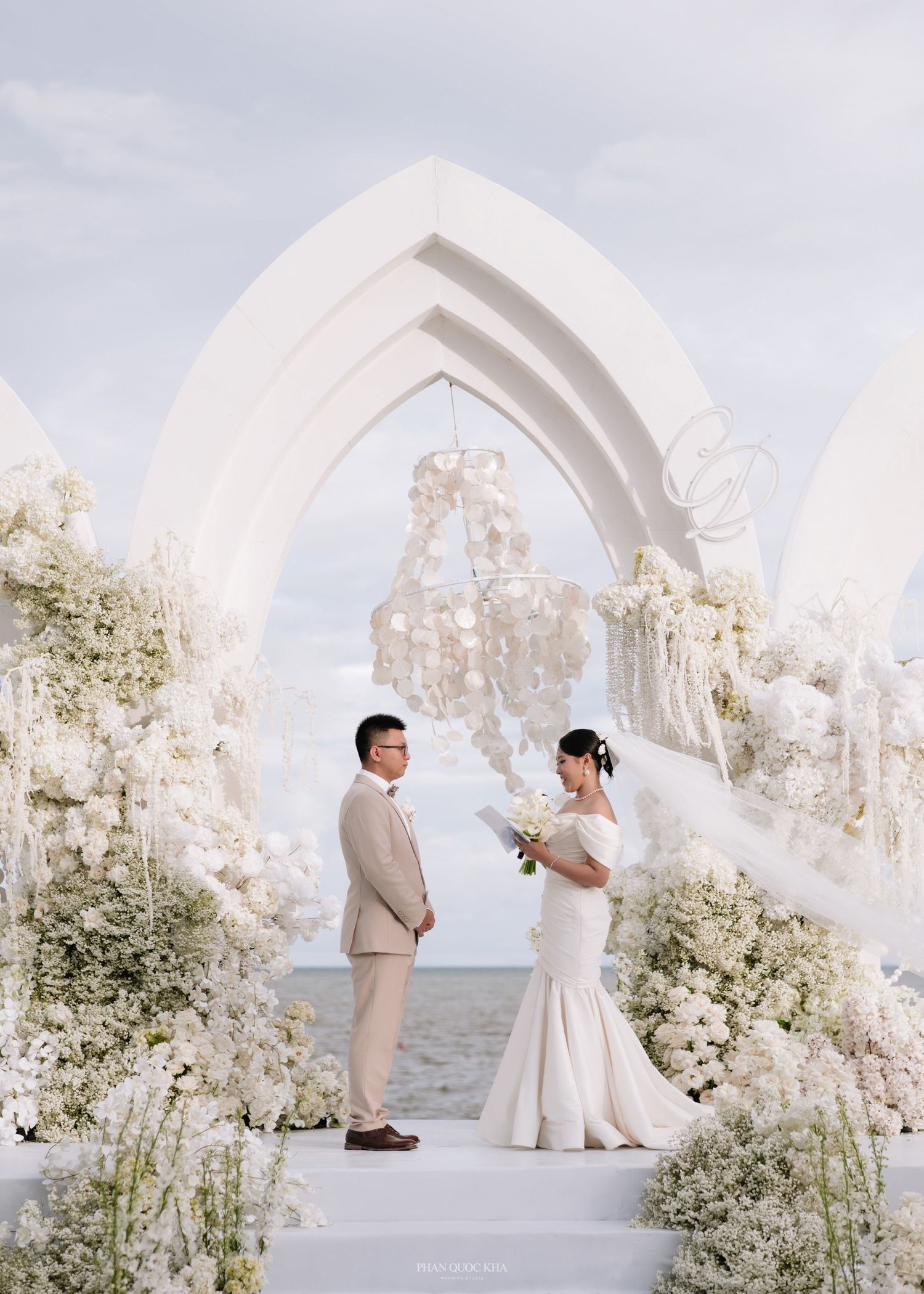 Couple exchanging vows at elegant outdoor beach wedding ceremony with white floral arches and chandelier backdrop.