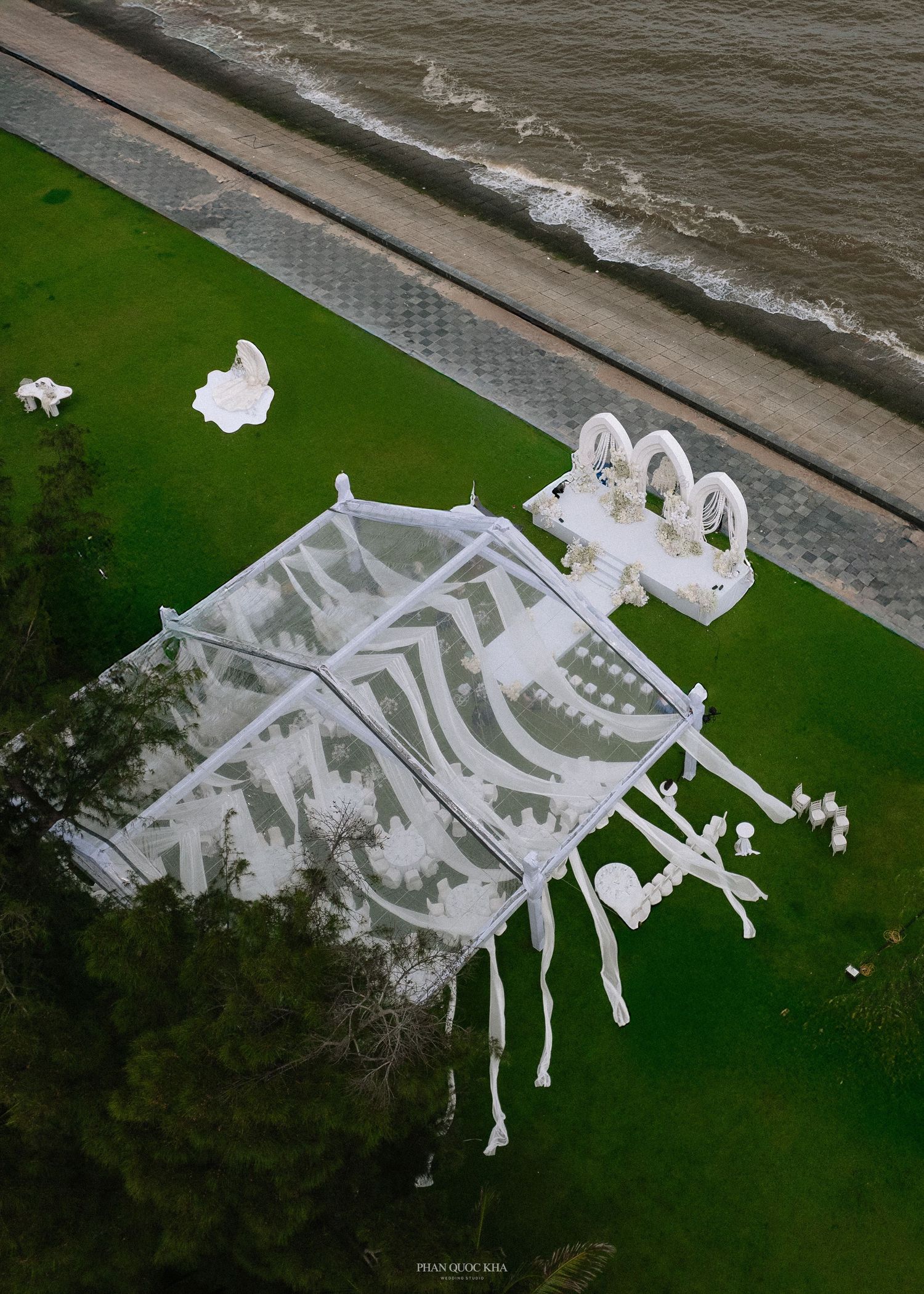 Aerial view of a white transparent event tent with decorative arches on green lawn near a beach.