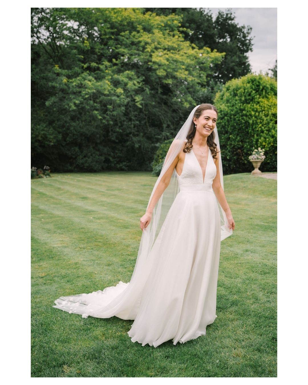 Bride in elegant white V-neck wedding gown with flowing train and veil, smiling outdoors on lush green lawn.