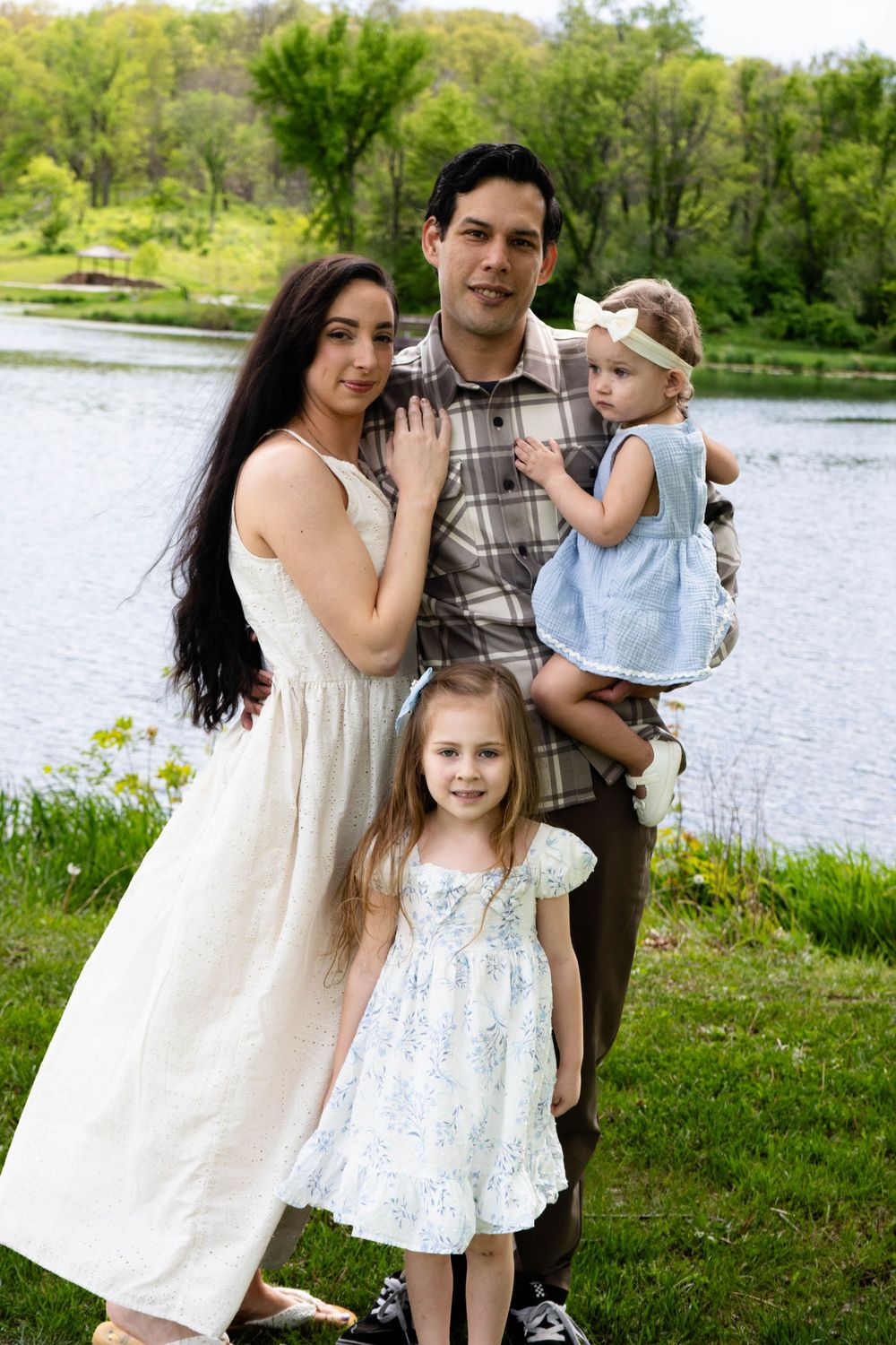 Family of four posing outdoors near a lake, parents with two young daughters in summer dresses surrounded by green trees.