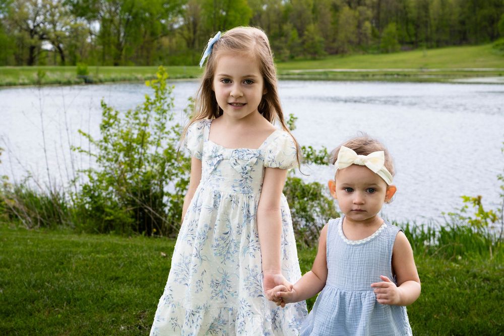Two young sisters in floral and blue dresses hold hands by a peaceful pond surrounded by green trees.