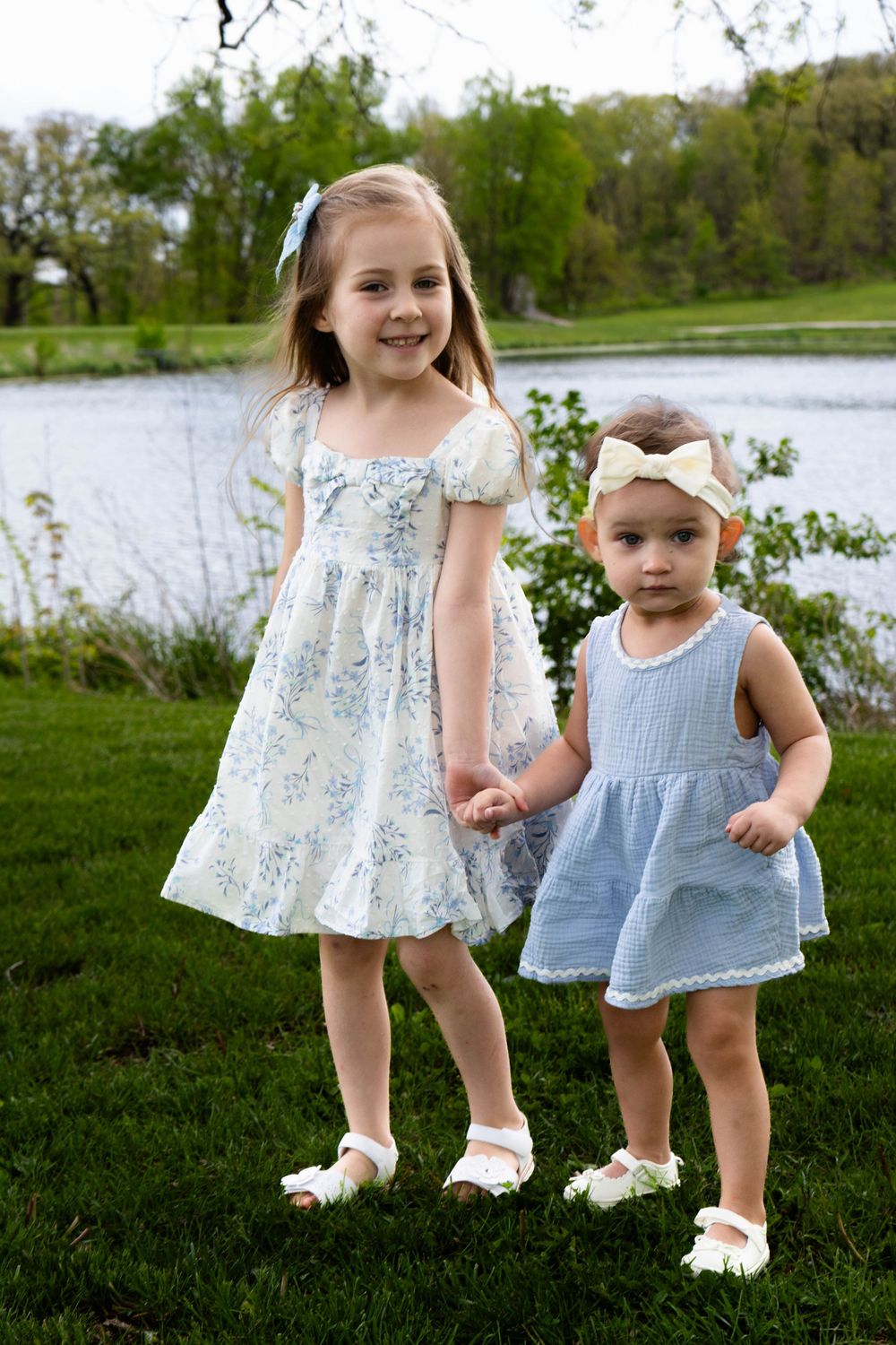 Two young sisters in matching blue and white dresses hold hands on green grass near a pond on a spring day.