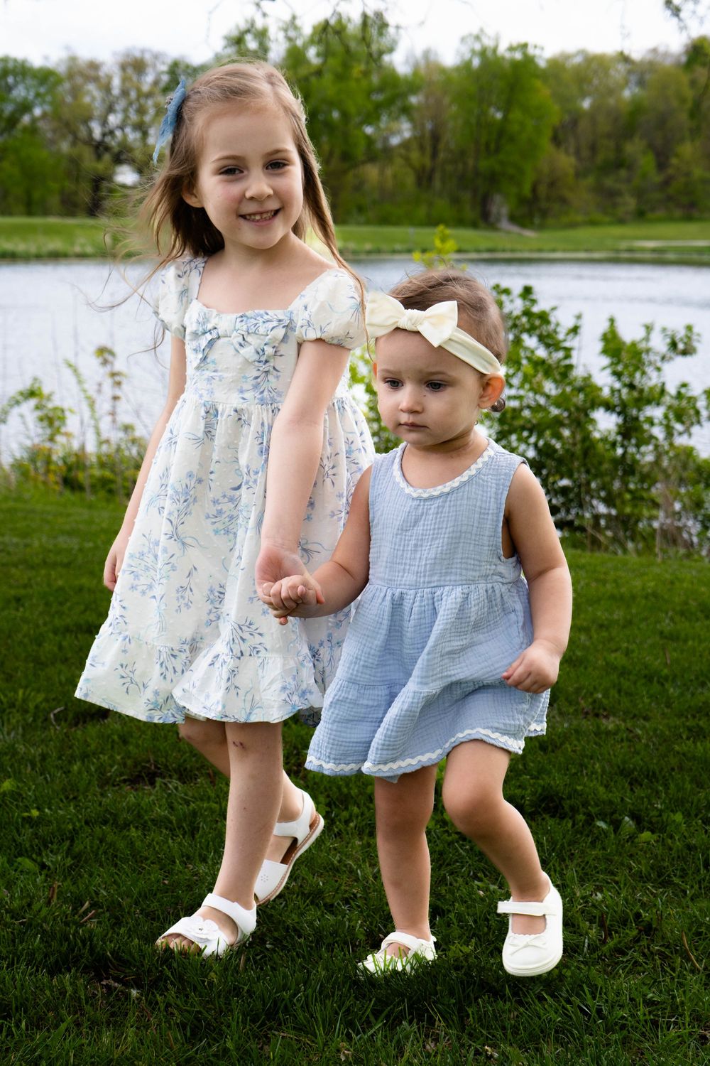 Two young sisters in matching blue dresses hold hands on green grass near a pond on a sunny spring day.