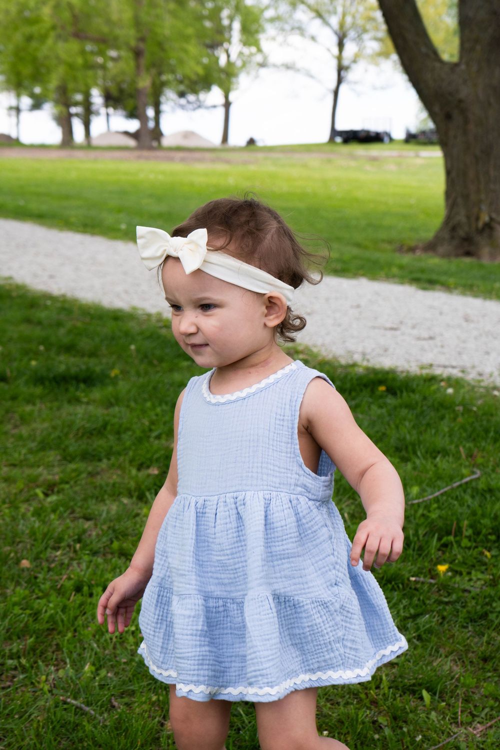 Toddler girl in blue dress and cream bow headband smiling outdoors in a green park setting.