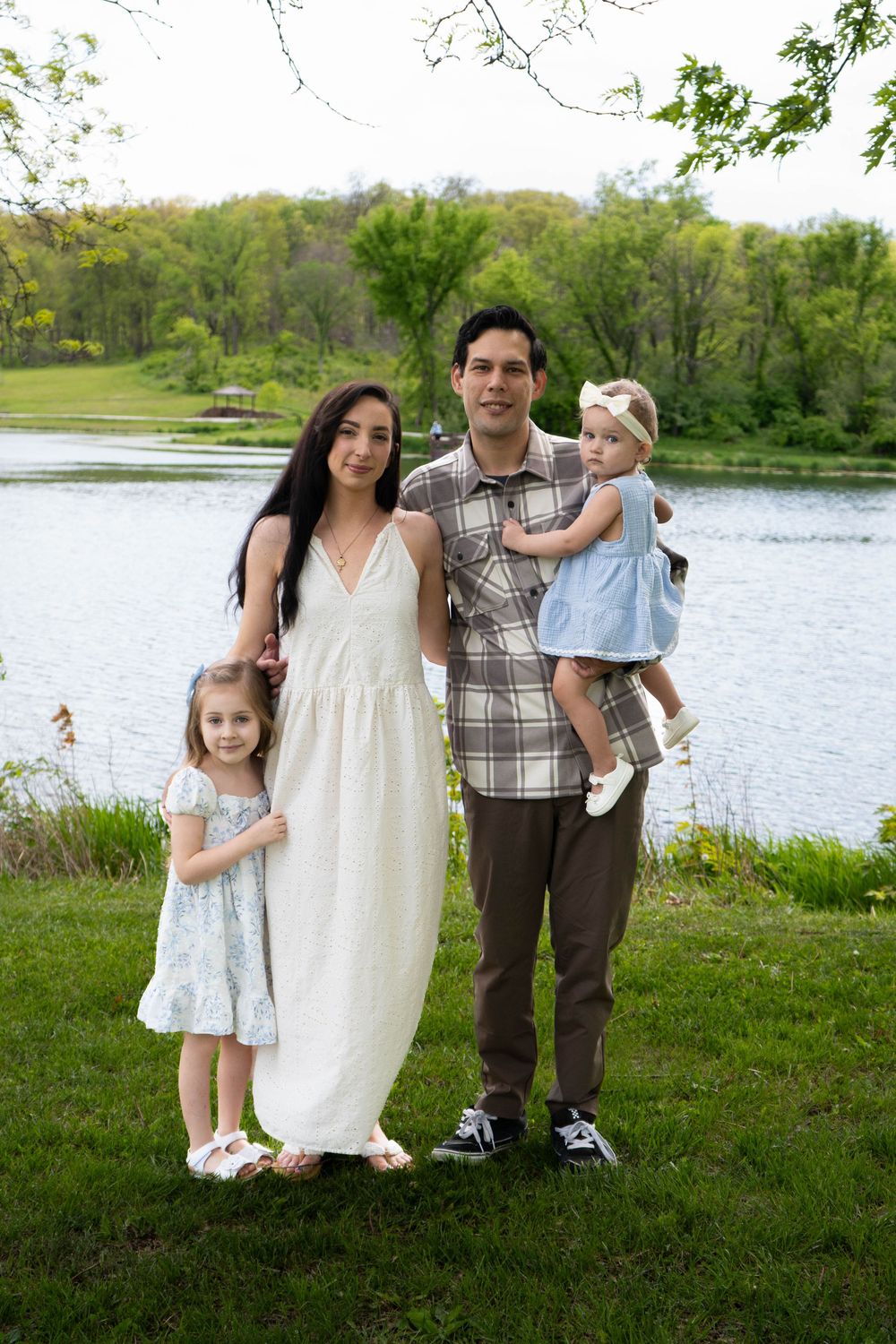 A family of four poses by a serene lake surrounded by lush green trees on a warm spring day.