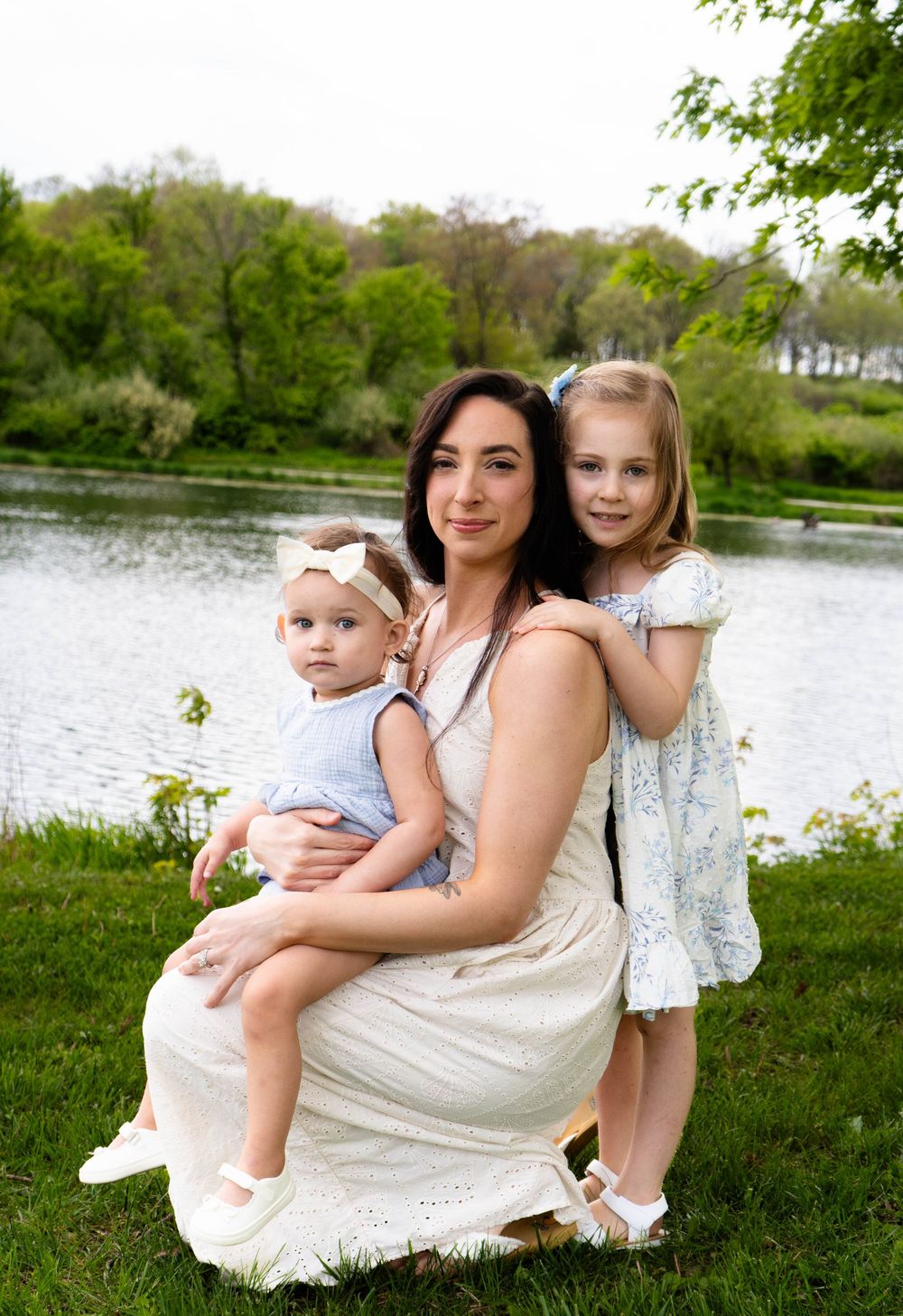 A mother sits outdoors by a lake holding her baby girl while her older daughter hugs her in a lush green park.
