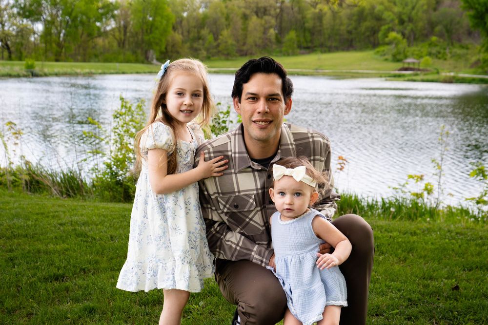 A man kneels with two young girls near a serene pond surrounded by lush green trees in a park setting.