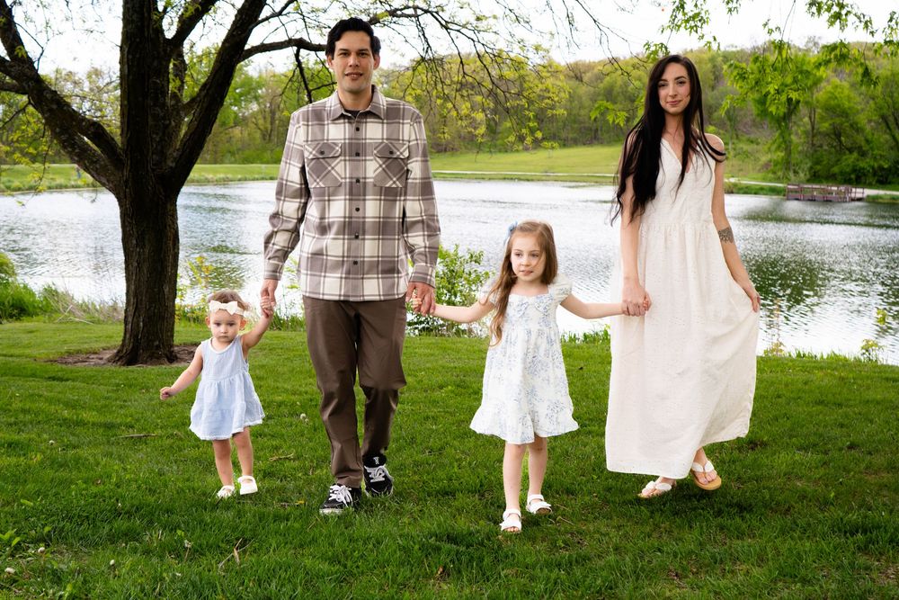 Family of four holding hands on green grass near a lake, two young girls in white dresses with parents on a sunny day.