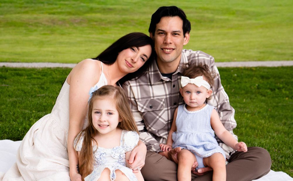 A happy young family with two little girls sitting on a blanket in a lush green park, smiling for a portrait.