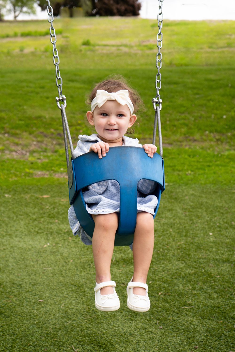 A smiling toddler girl with a white bow headband sits in a blue bucket swing at a playground.