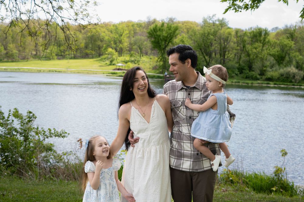 Happy family of four posing outdoors near a serene lake surrounded by lush green trees on a sunny day.