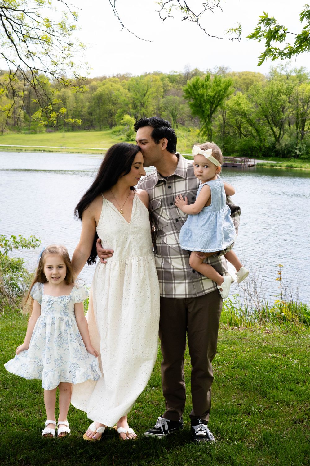 Family of four posing by a serene lake in a lush green park, mother in white dress, father holding toddler girl.