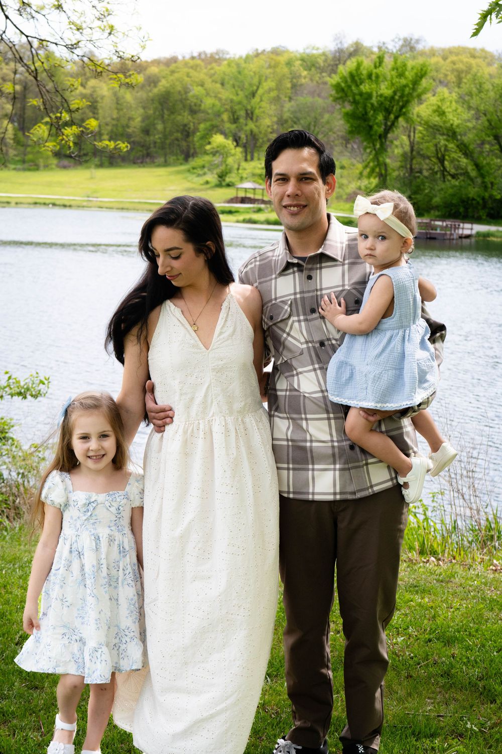 Family of four posing outdoors near a serene lake surrounded by lush green trees on a spring day.