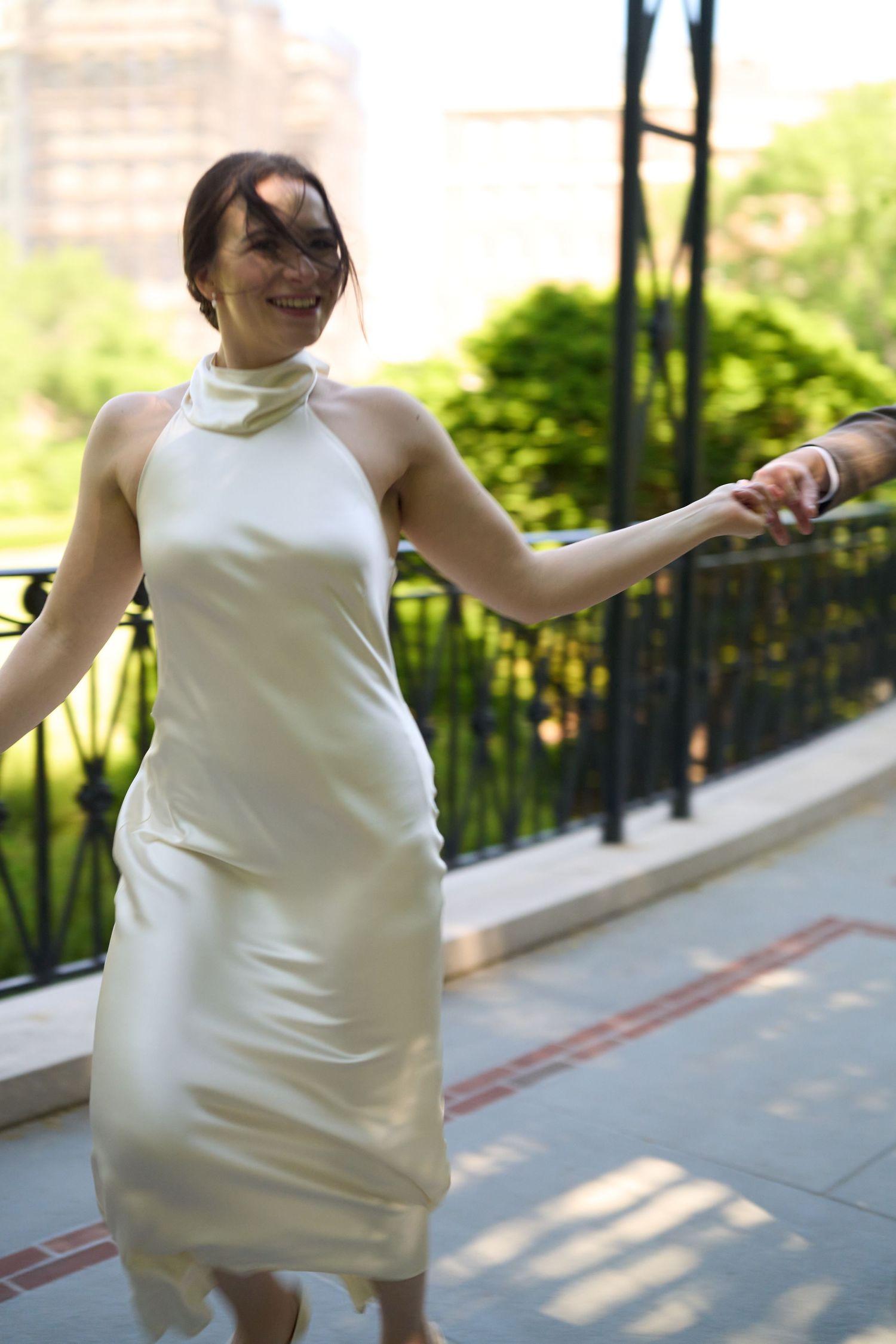 A joyful woman in an elegant white halter dress twirls outdoors on a sunny day near a black iron railing.