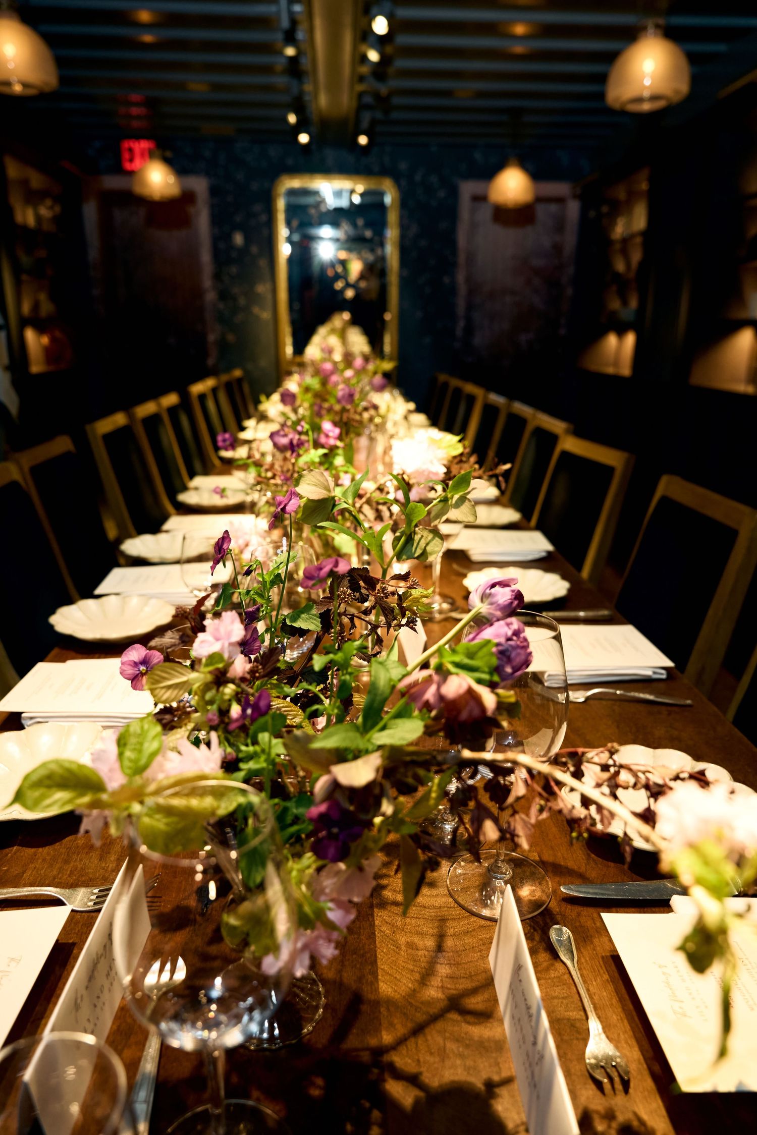 Elegant long dining table set with floral centerpieces, white plates, and candles in a dimly lit restaurant.
