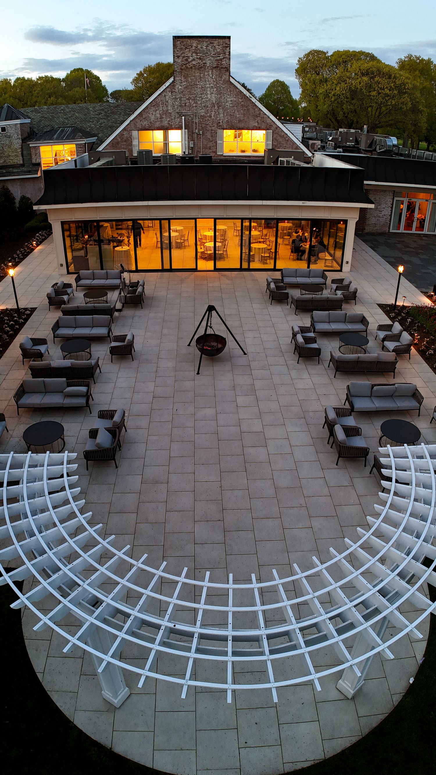 Aerial view of an elegant outdoor patio with white pergola, lounge seating, and a glowing glass building at dusk.
