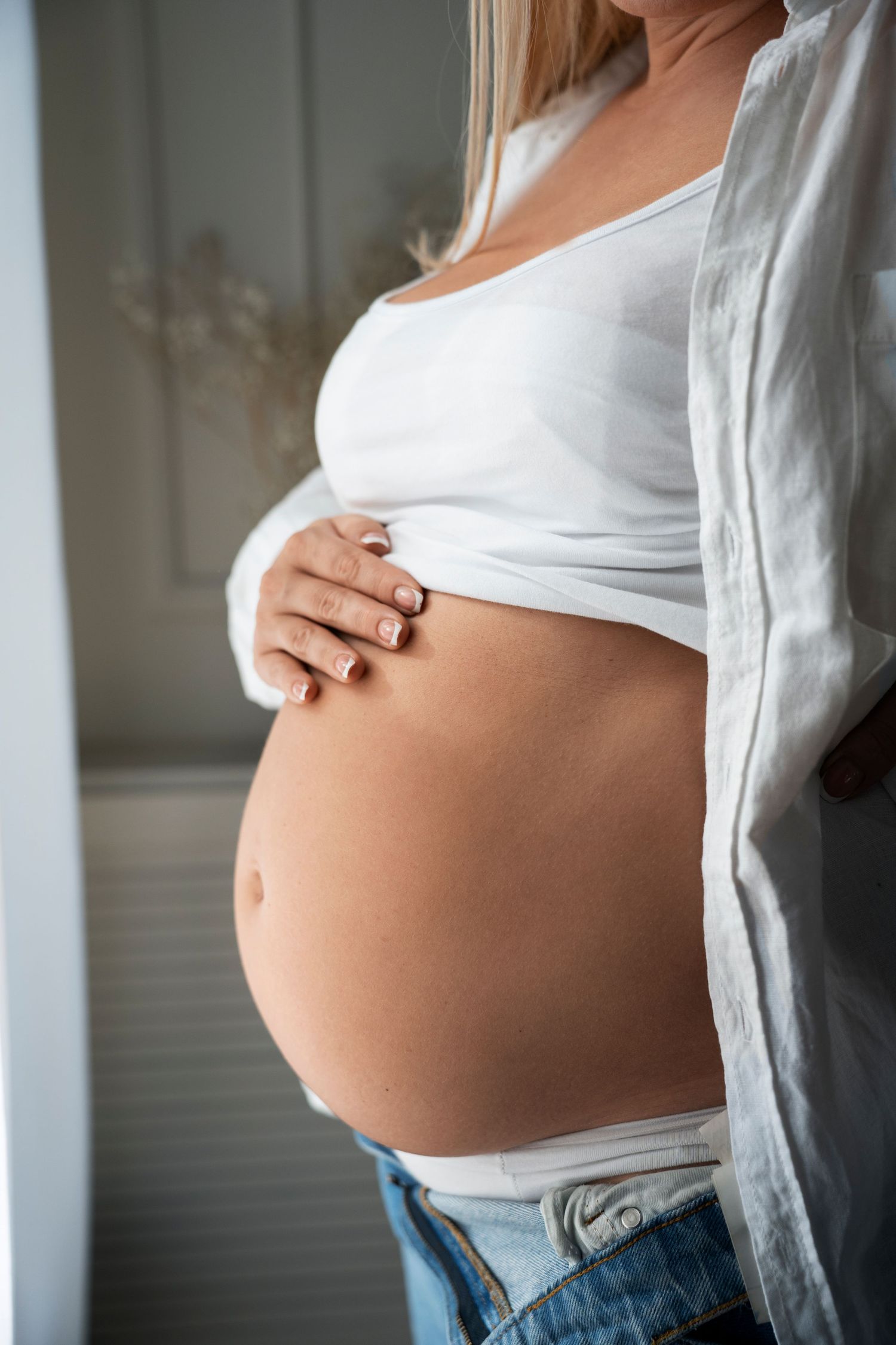 Une femme enceinte, vêtue d'un haut blanc et d'un jean, caresse doucement son ventre nu, debout près d'une fenêtre lumineuse.