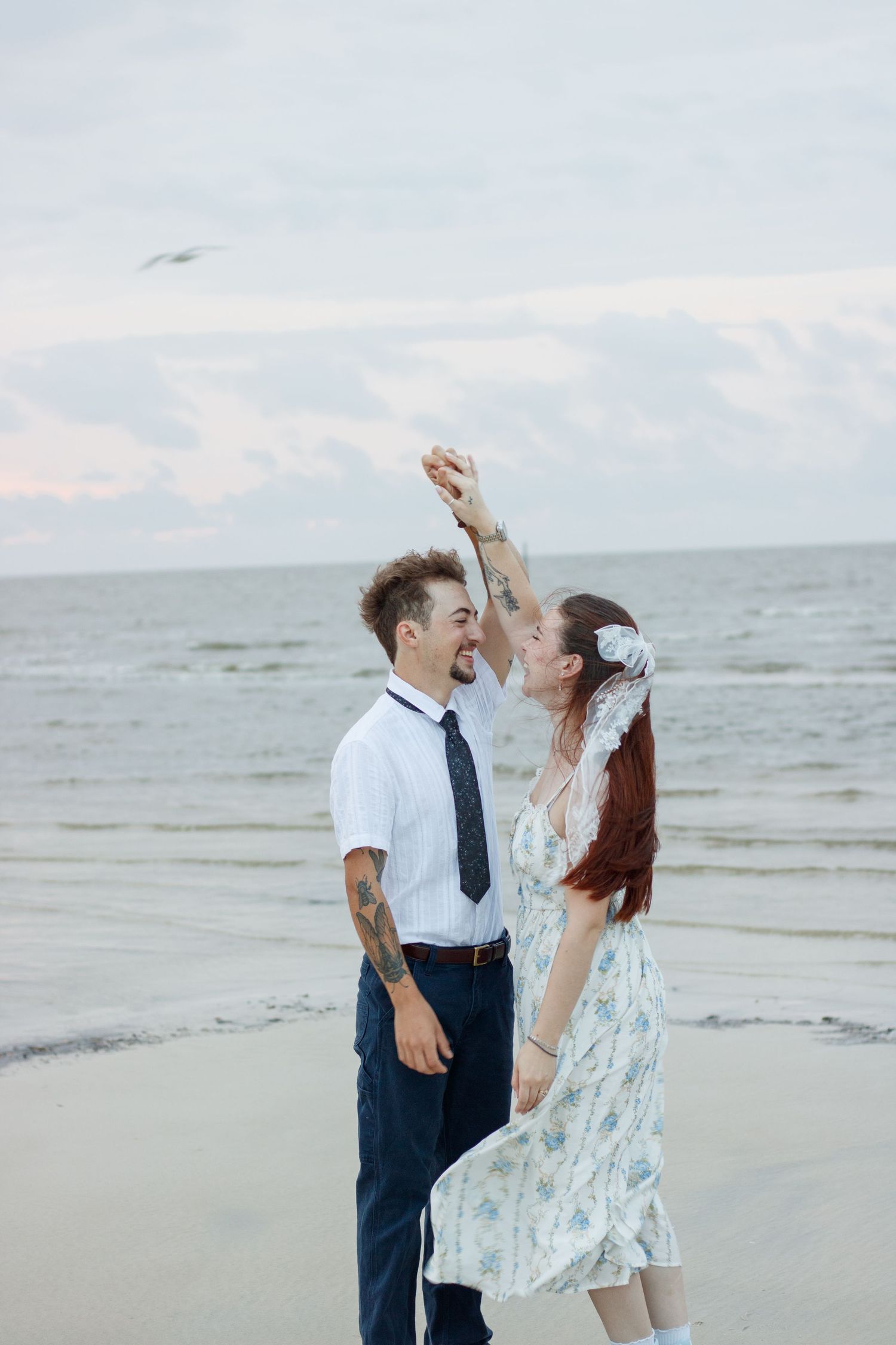 Couple laughing and dancing on a beach at dusk as the man twirls the woman in a floral dress.