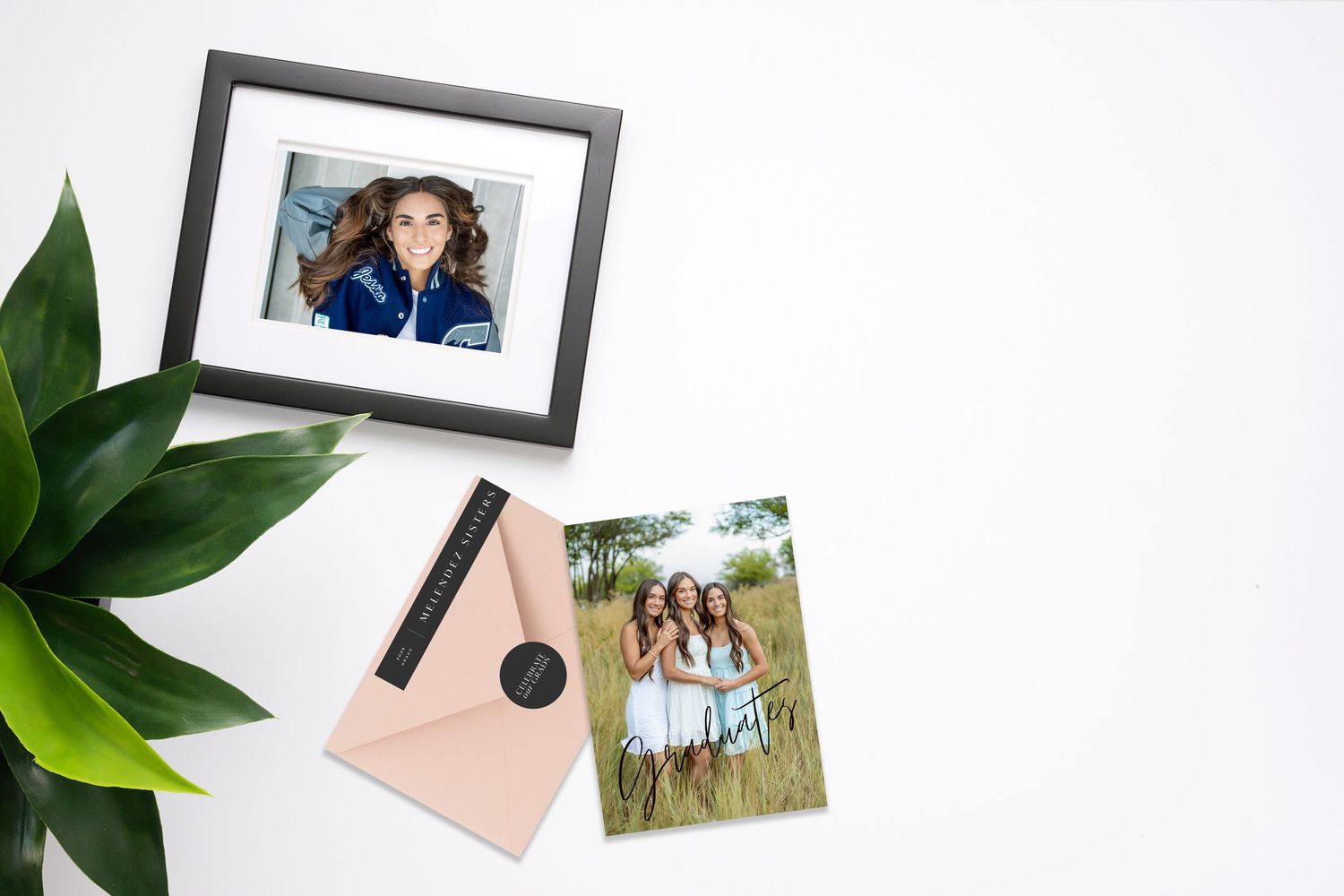 Framed portrait photo display with loose prints, pink envelope, and green plant on white surface.