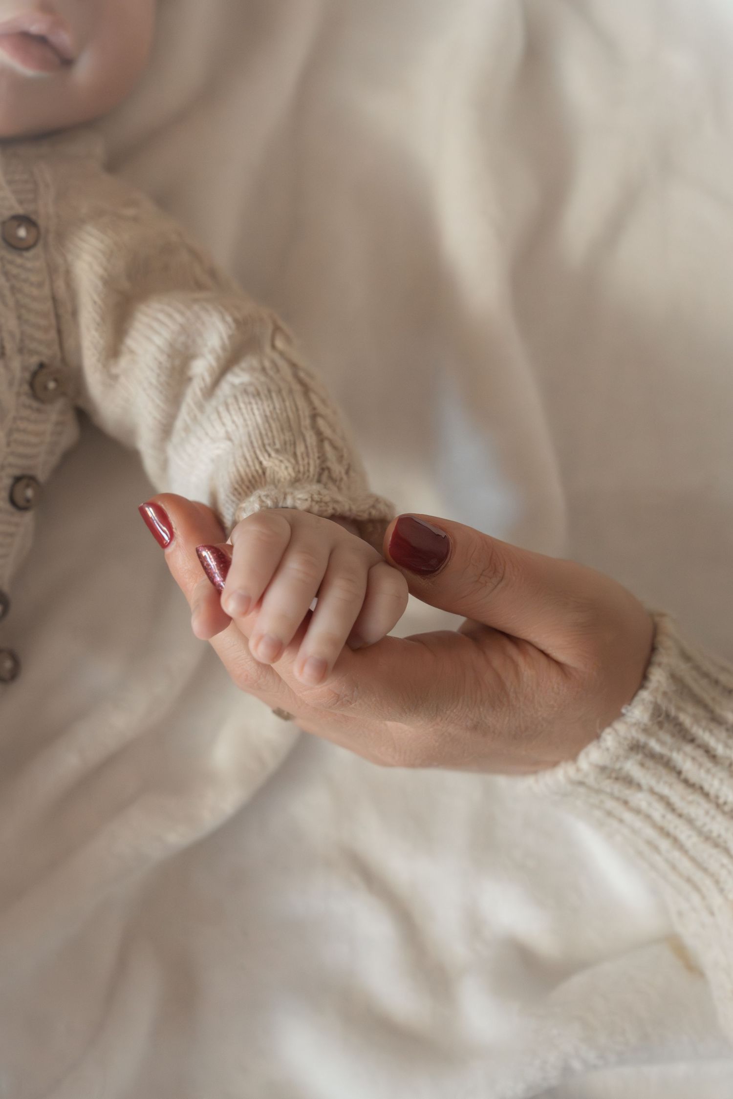 A mother with red nails gently holds her newborn baby's tiny hand, both wearing cozy cream knit sweaters.
