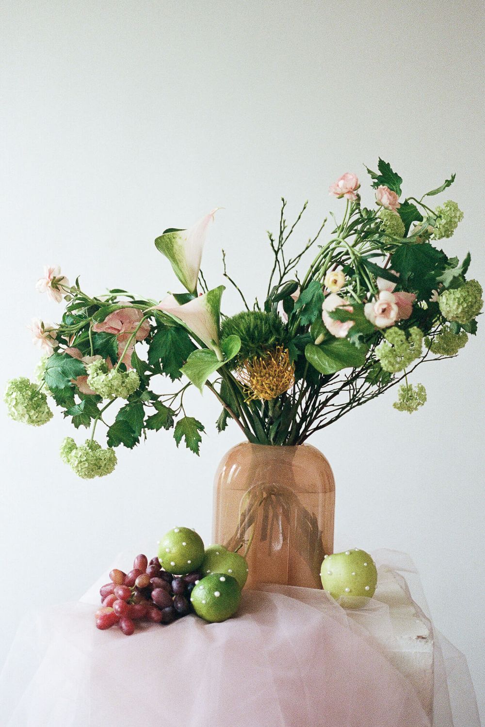 A floral arrangement in a amber vase with pink lilies and green blooms beside fresh grapes and green apples on a pink cloth.