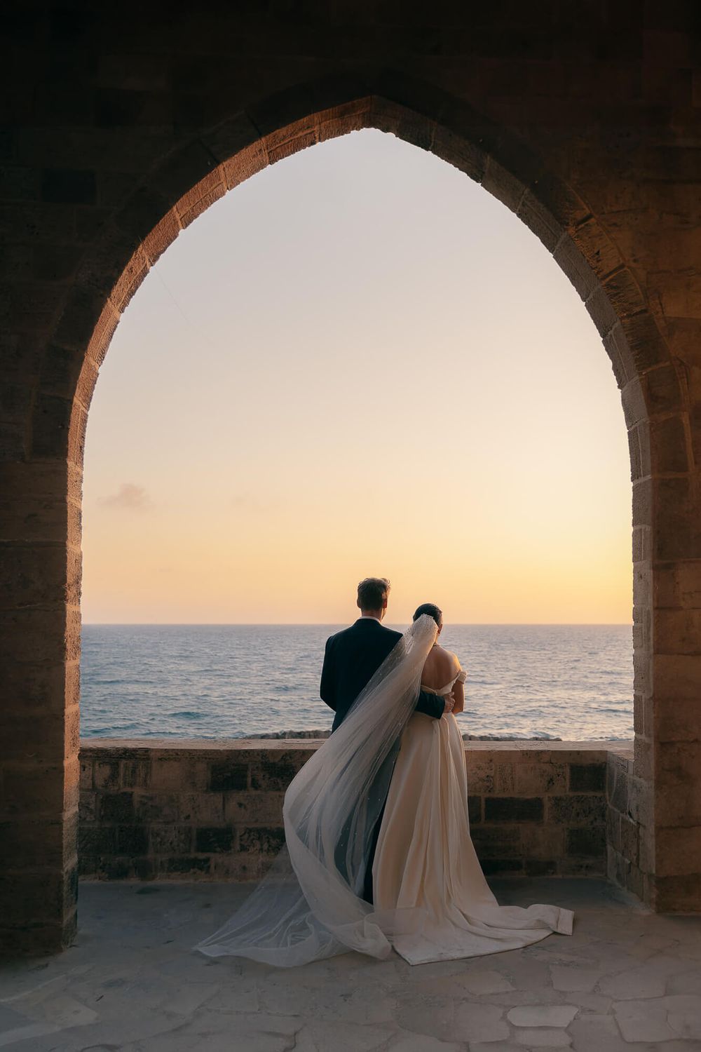 Bride and groom stand under stone arch overlooking ocean sunset at romantic seaside wedding venue.