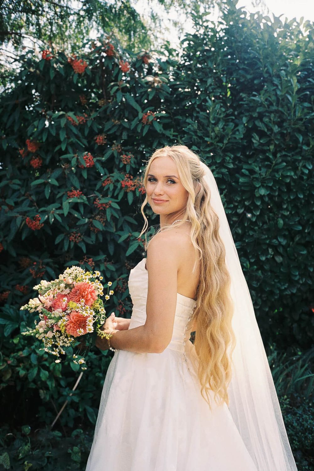 Smiling bride with long blonde hair holding a colorful bouquet in a white strapless gown, standing before lush green foliage.