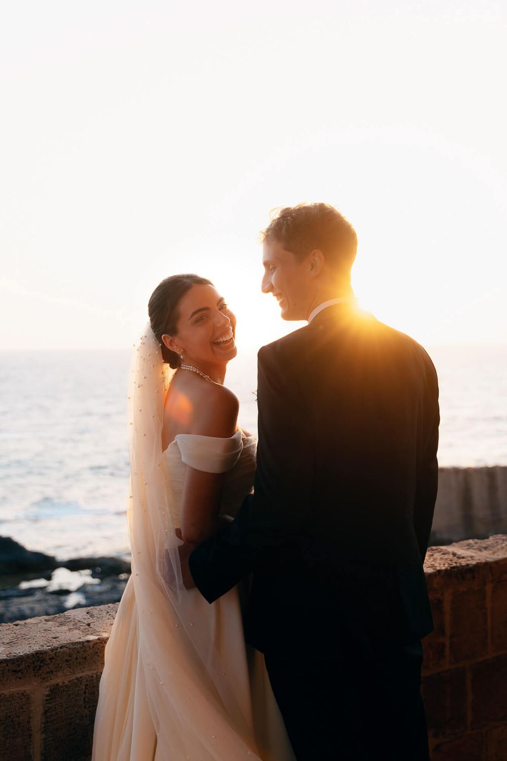 Bride and groom share a romantic sunset moment by the ocean, bride smiling in white off-shoulder gown with veil.