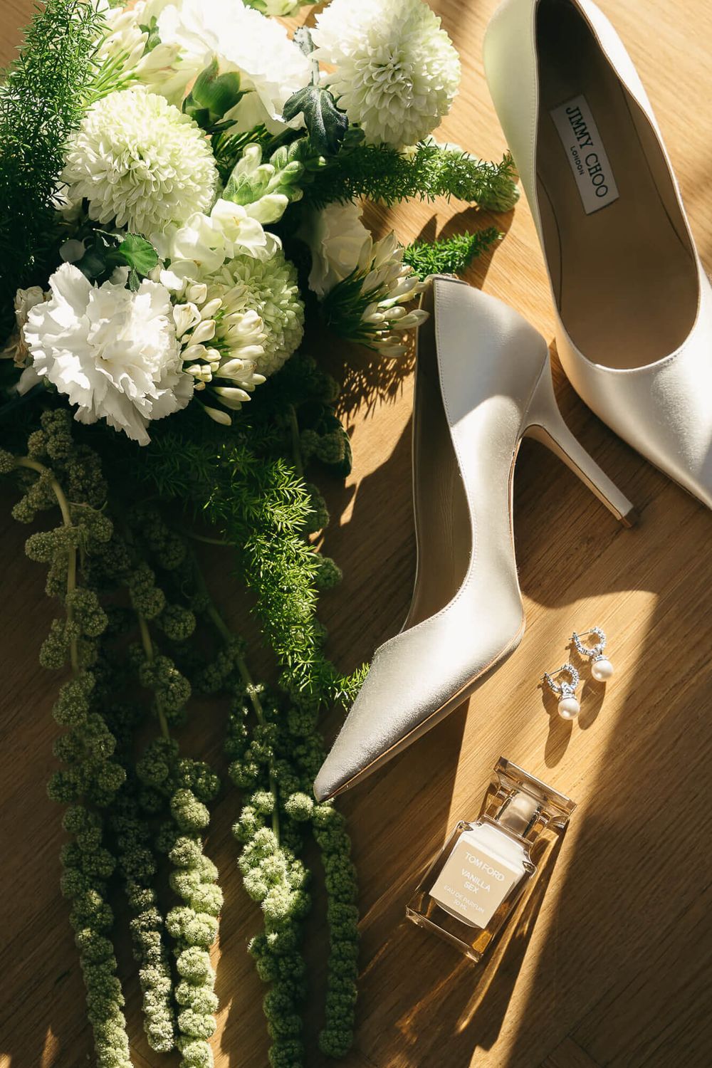 White Jimmy Choo bridal heels beside a white floral bouquet, pearl earrings, and perfume on a wooden surface.