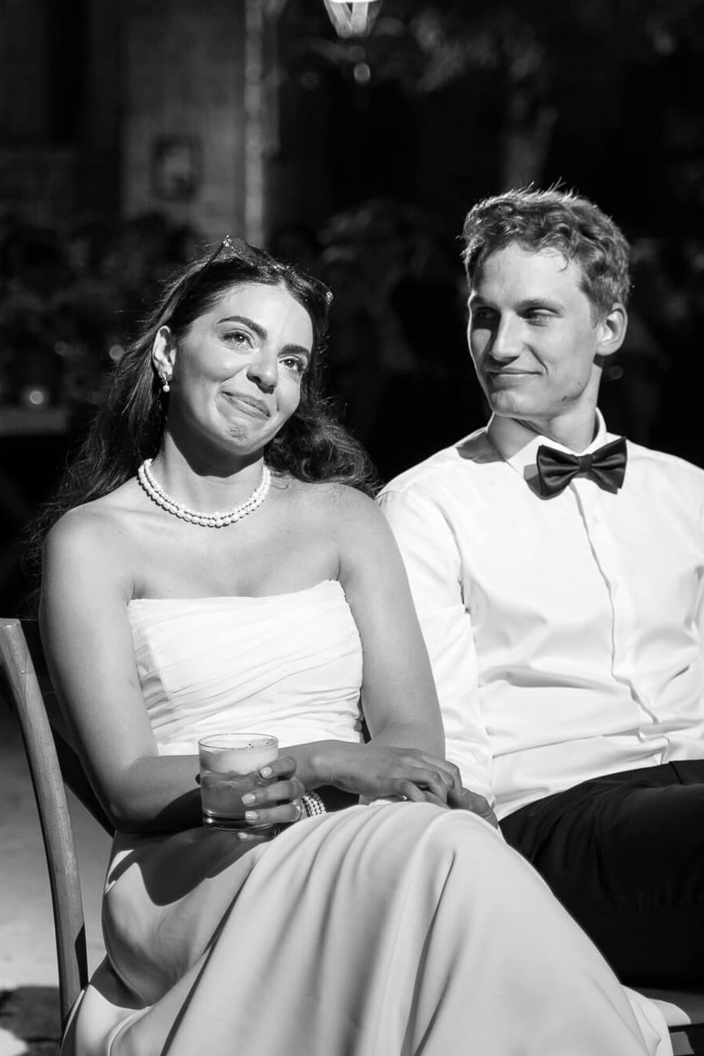 Black and white photo of a bride and groom smiling at their wedding reception, bride in strapless gown with pearl necklace.