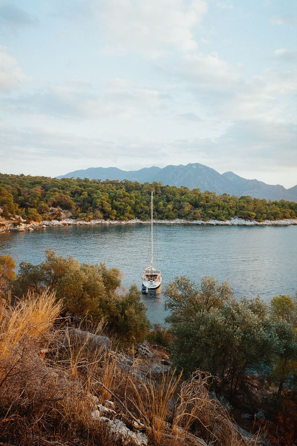 A sailboat anchored in a calm Mediterranean cove surrounded by lush green hills and mountains under a cloudy sky.