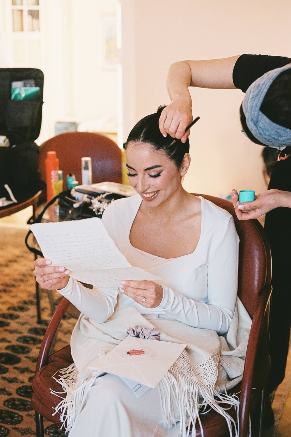 Smiling bride reads a letter while getting her hair styled in a warmly lit room before her wedding.