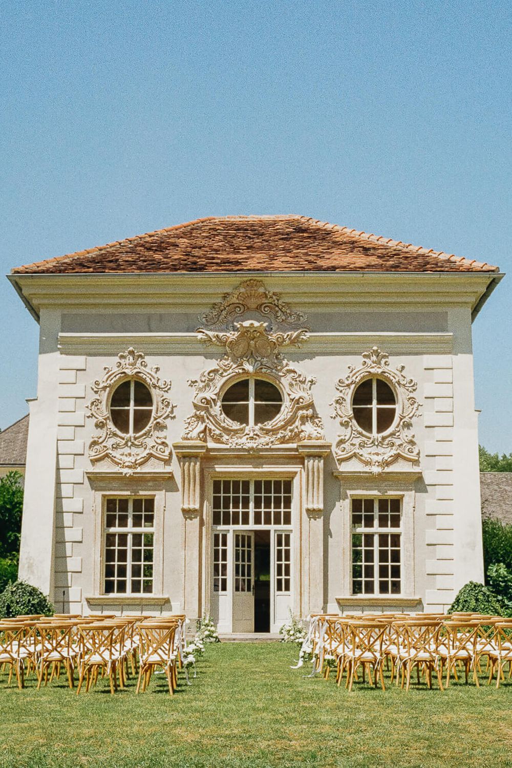 Ornate white baroque-style wedding venue with wooden chairs arranged on lawn under clear blue sky.