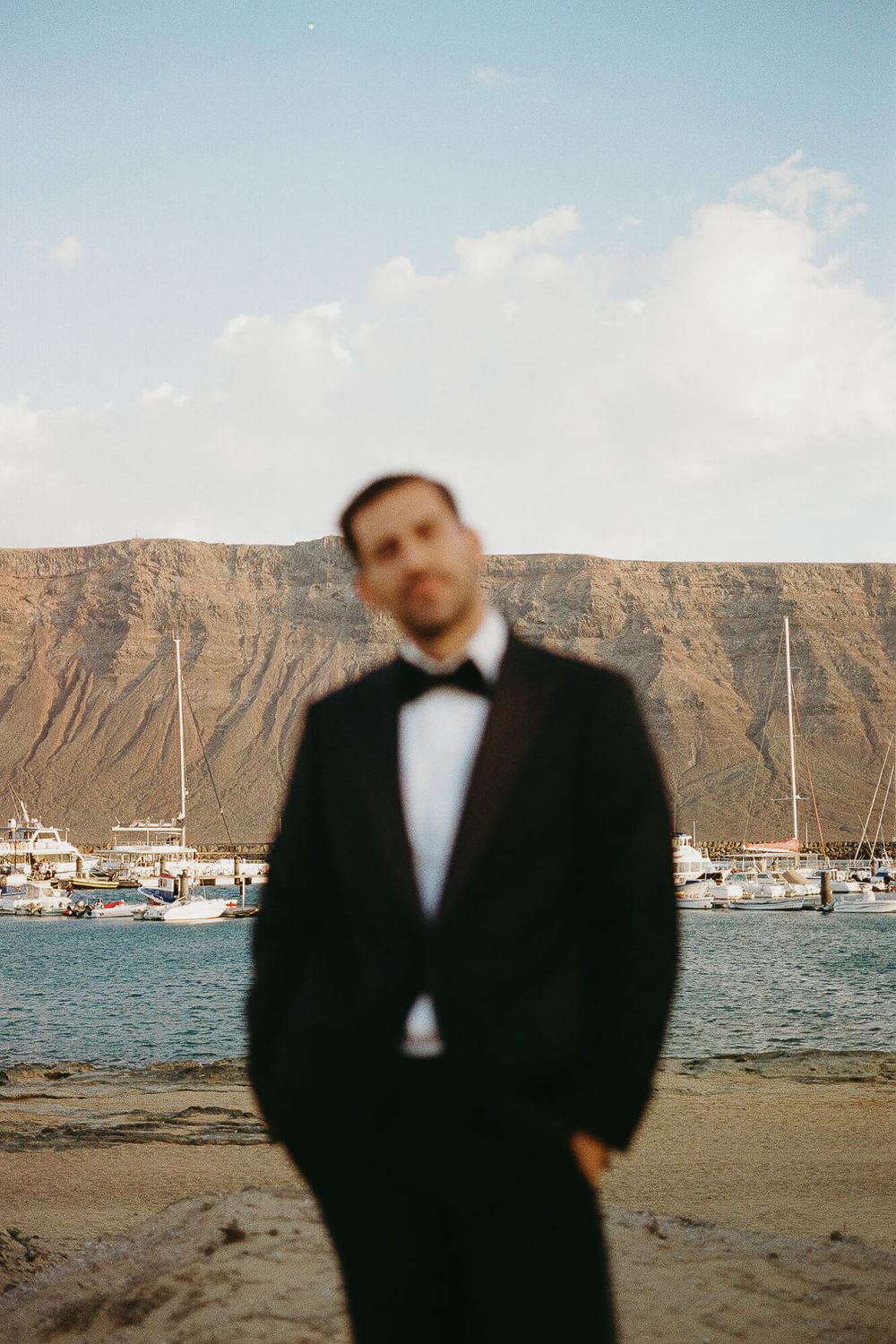 A blurred groom in a black tuxedo stands on a sandy beach with boats and a rocky cliff in the background.