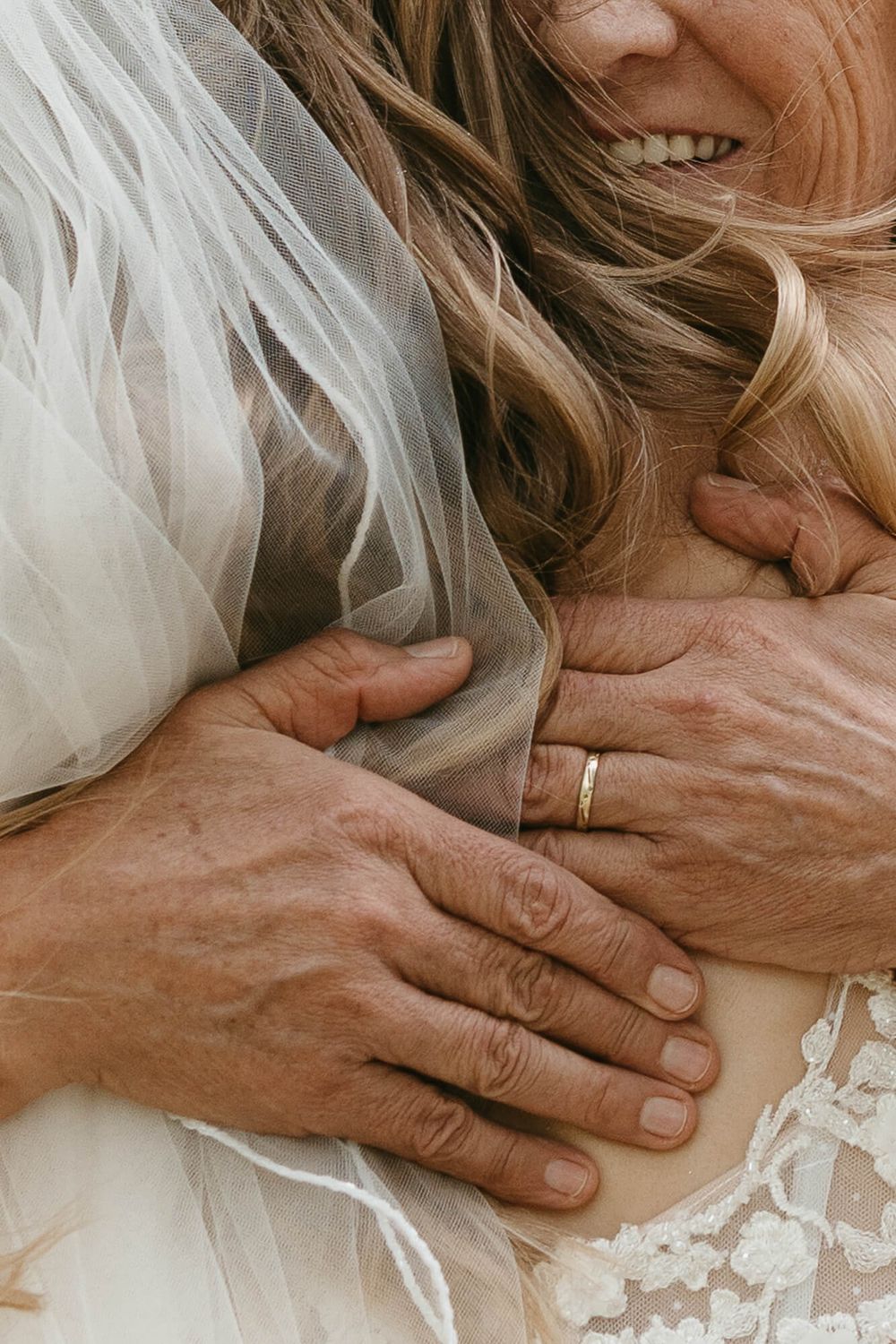Close-up of groom's hands with wedding band embracing bride in lace dress and veil on wedding day.