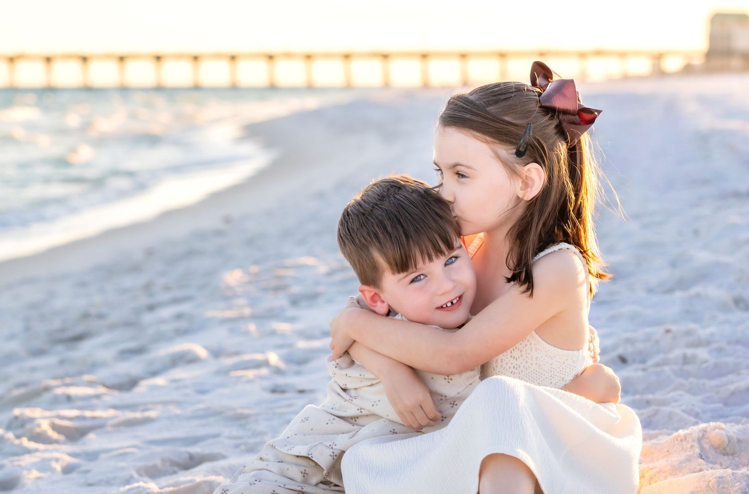 Two young children share a sweet hug on a sandy beach at sunset, wearing white outfits.