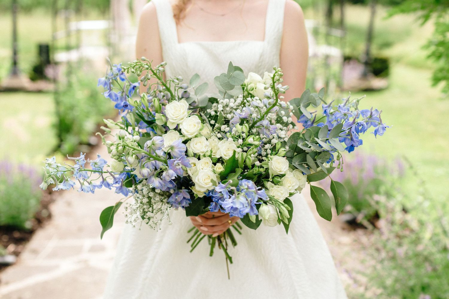 Bride holding a lush blue and white wedding bouquet with delphiniums, roses, and greenery outdoors.