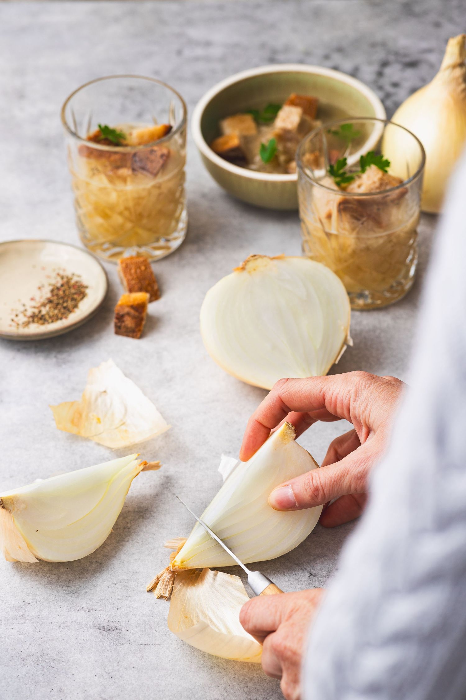 Hands cutting a white onion on a marble surface with French onion soup in glasses and a bowl nearby.