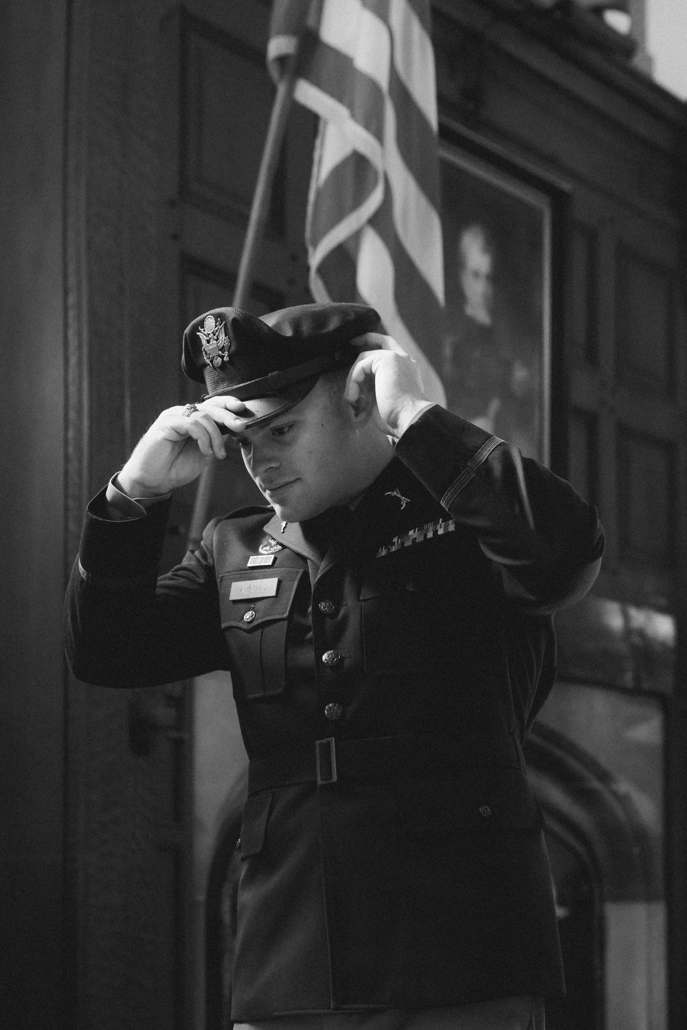 Black and white photo of a military officer in dress uniform adjusting his cap, with an American flag in the background.