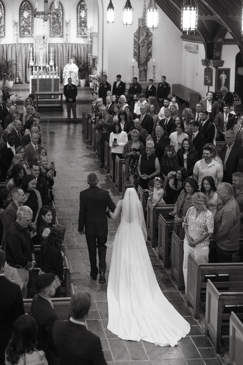 Bride walks down church aisle with escort in black and white wedding ceremony photo with guests in pews.