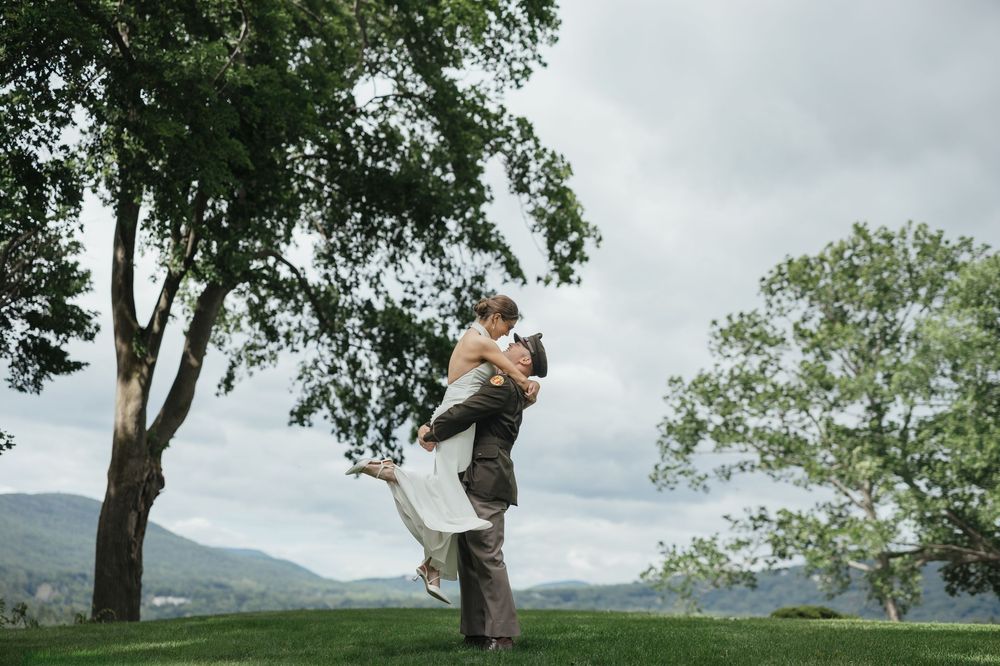 Groom lifts bride in wedding attire on lush green hillside with mountain views and trees under cloudy sky.