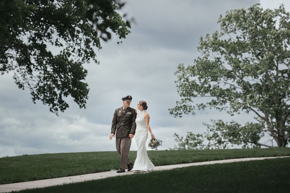 Military groom in uniform walks with bride in white gown on green lawn under cloudy sky at outdoor wedding.