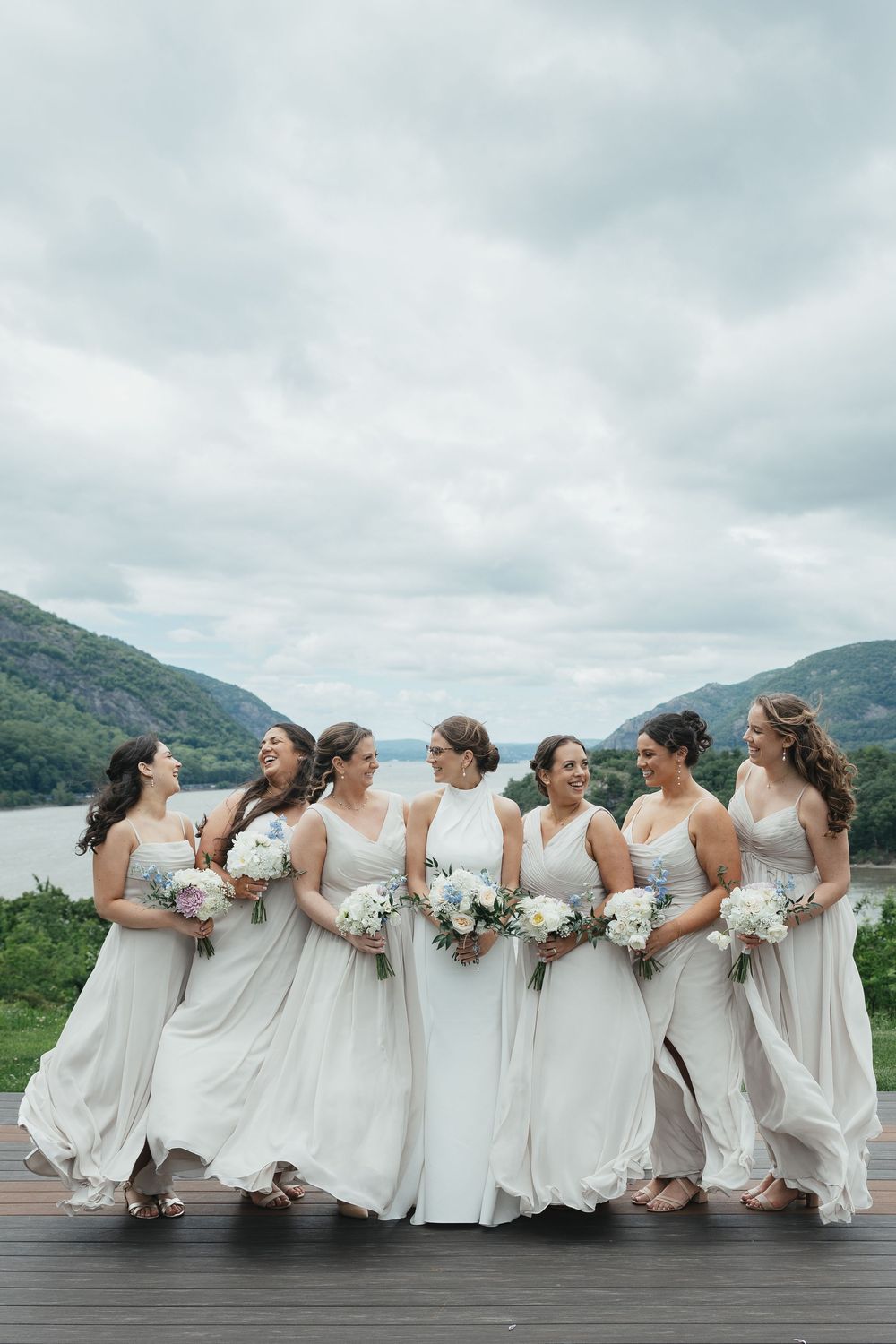 Bride in white gown with bridesmaids in champagne dresses holding bouquets on deck with mountain lake view.