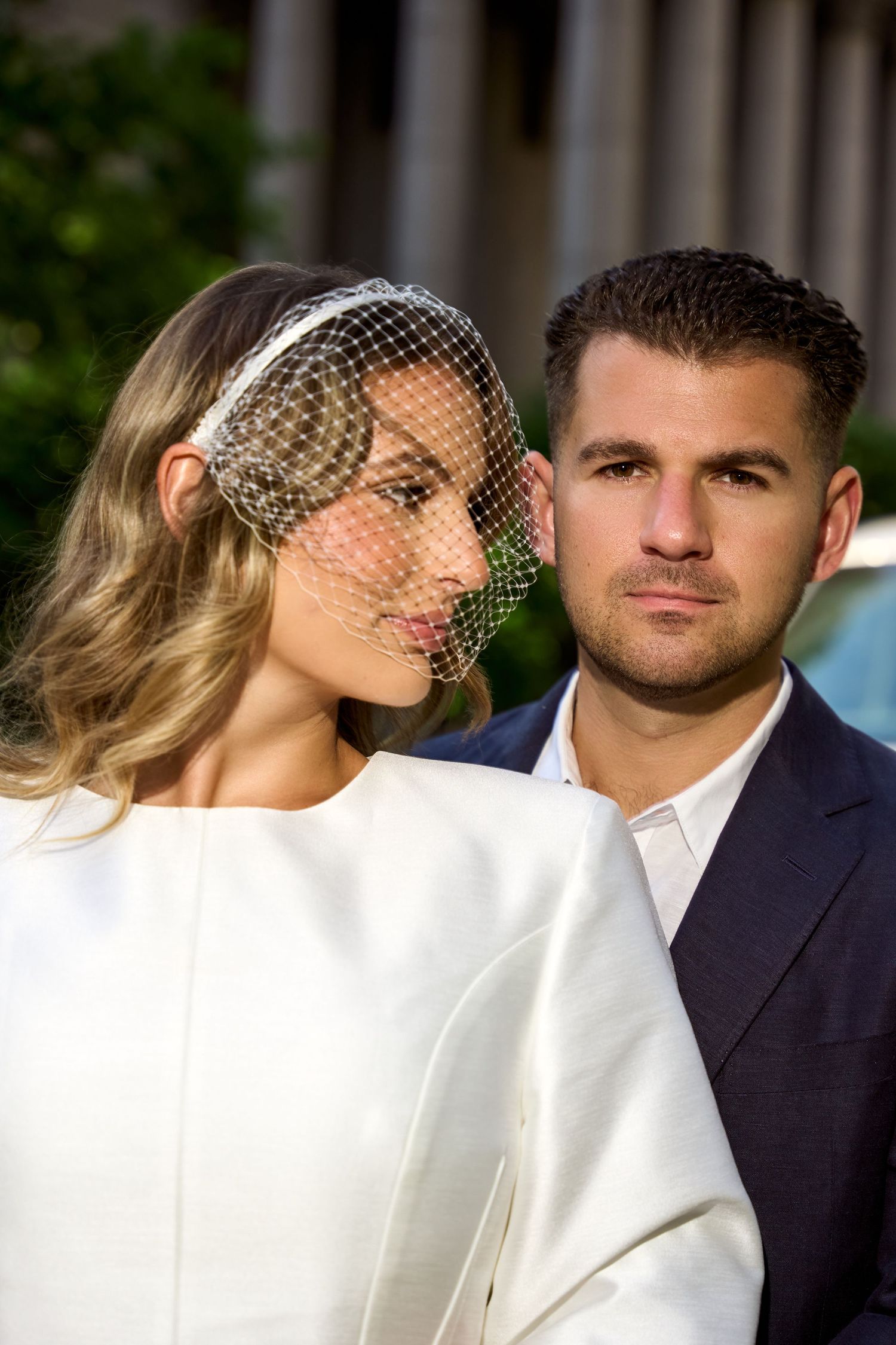 Elegant bride in white dress with birdcage veil poses with groom in navy suit at outdoor wedding ceremony.