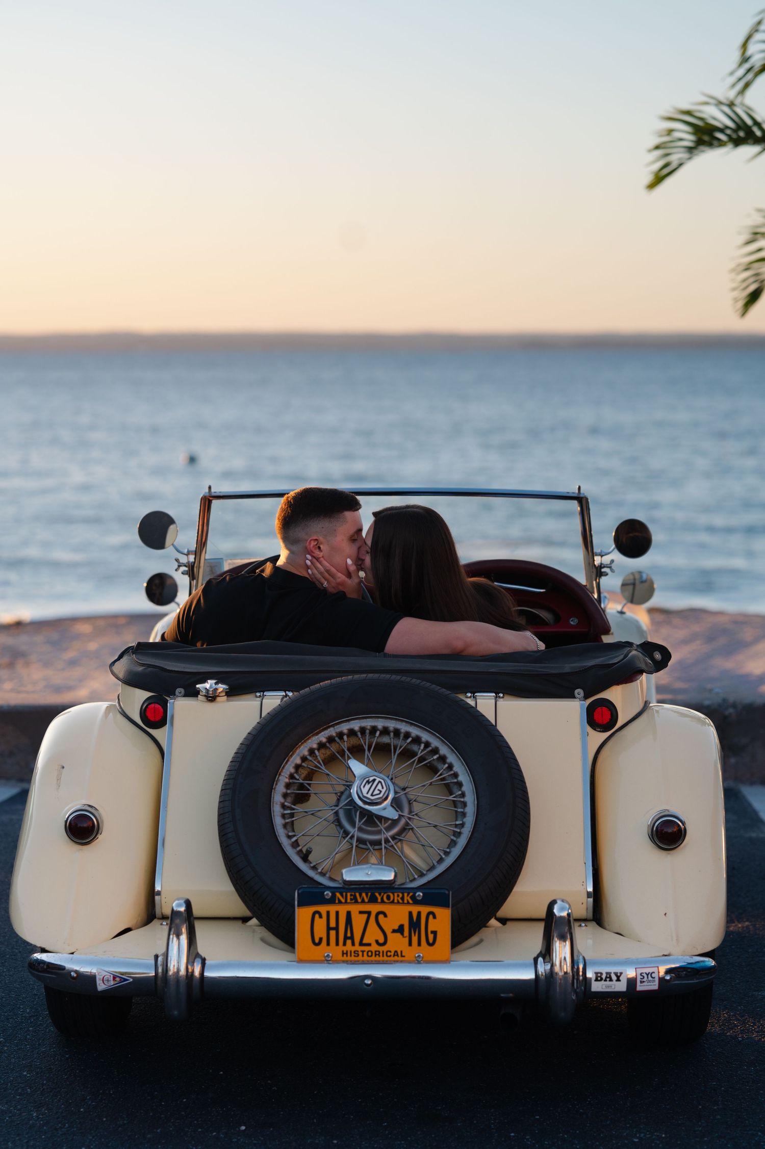 Couple kissing in a vintage cream convertible car with license plate CHAZS-MG at sunset by the ocean.