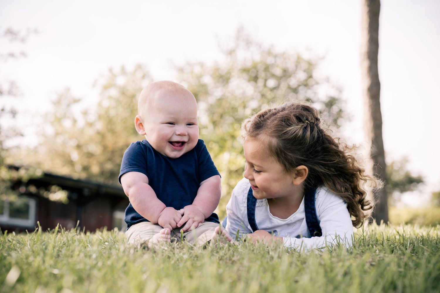 A laughing baby and young girl playing together on green grass outdoors in a sunny park setting.