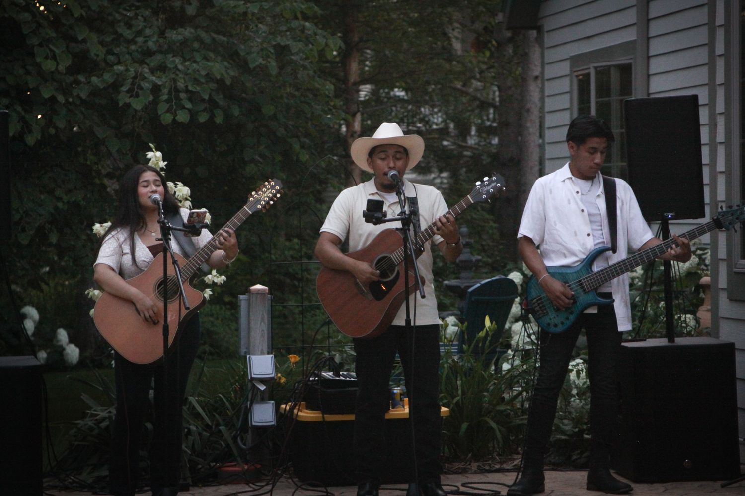 Three musicians perform outdoors, two playing acoustic guitars and one electric guitar, with a vocalist in a white cowboy hat.